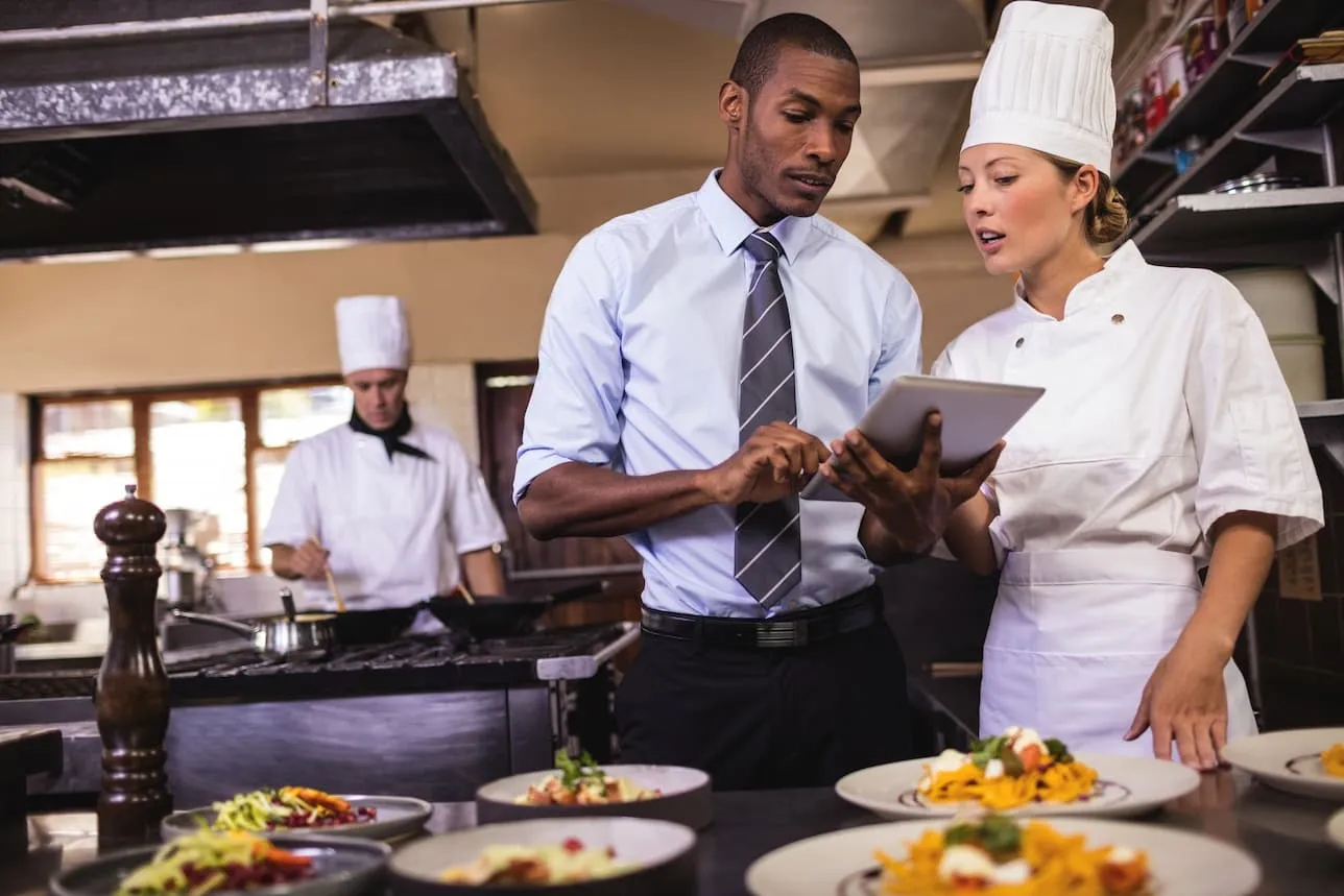 UK Food Allergen Regulations & Legal Compliance Framework: Complete Guide for Food Businesses 4 A manager and chef discuss food safety training in a commercial kitchen with prepared dishes on the counter.