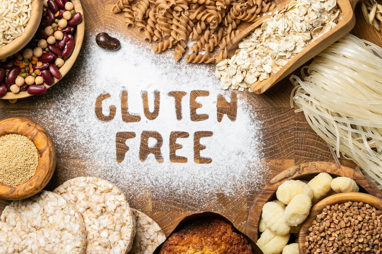 Various gluten-free grains and foods arranged around the words 'GLUTEN FREE' written in flour on a wooden surface.