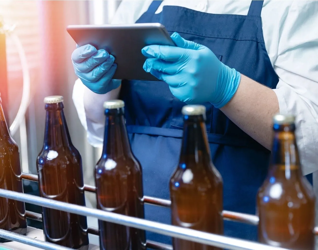 Allergen Recall Procedures UK: FSA Notification Requirements and Effective Incident Management 3 Person wearing blue gloves and apron inspecting brown bottles on a production line using a tablet.