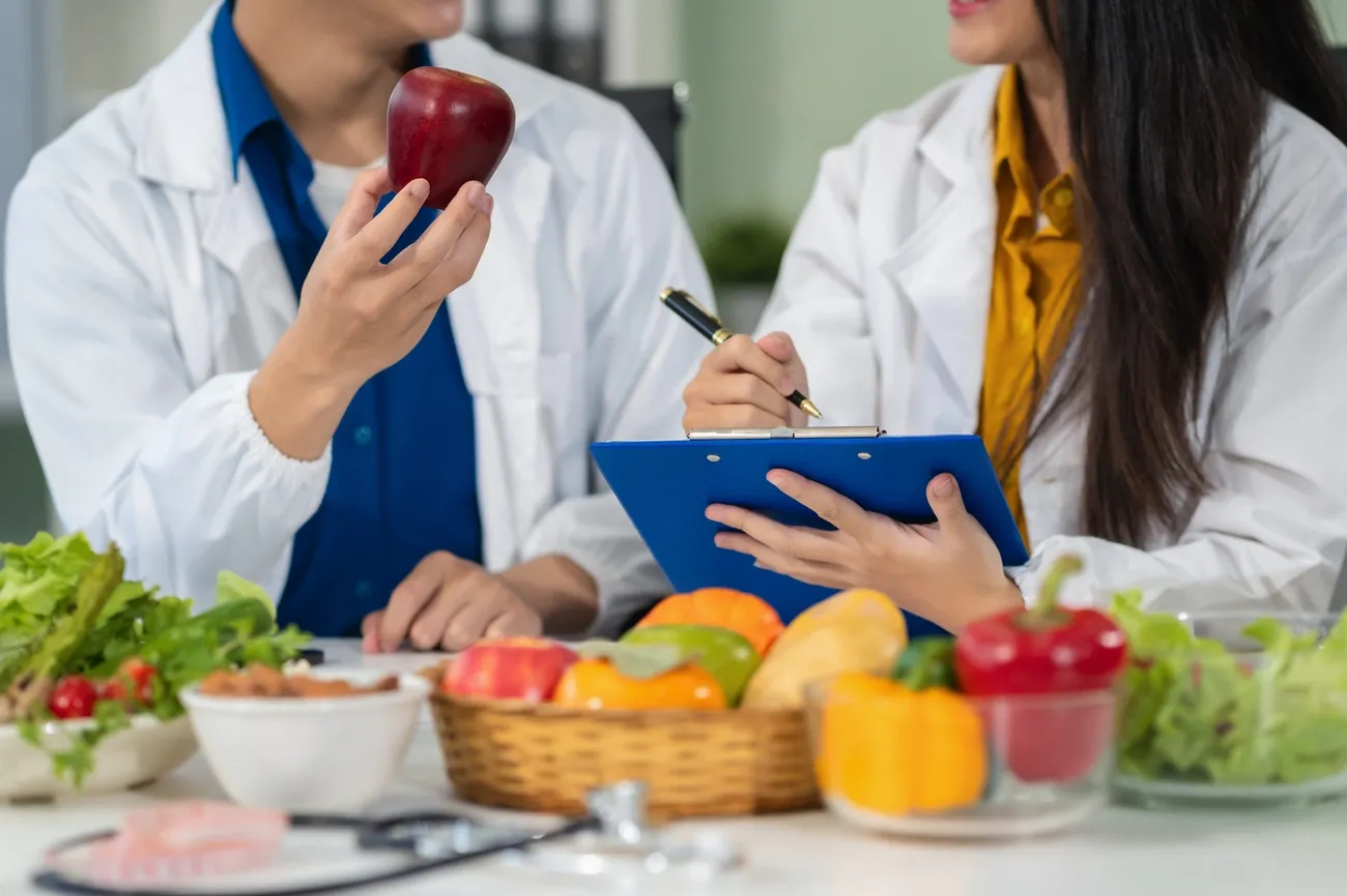 Two food scientists in lab coats discussing food allergen management with fresh produce and a clipboard.
