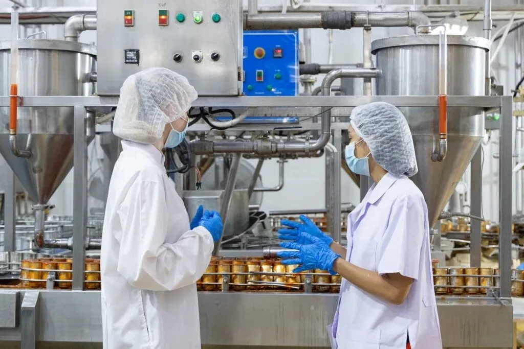 Two food safety workers in protective gear discussing near machinery in a food processing plant with canned products.