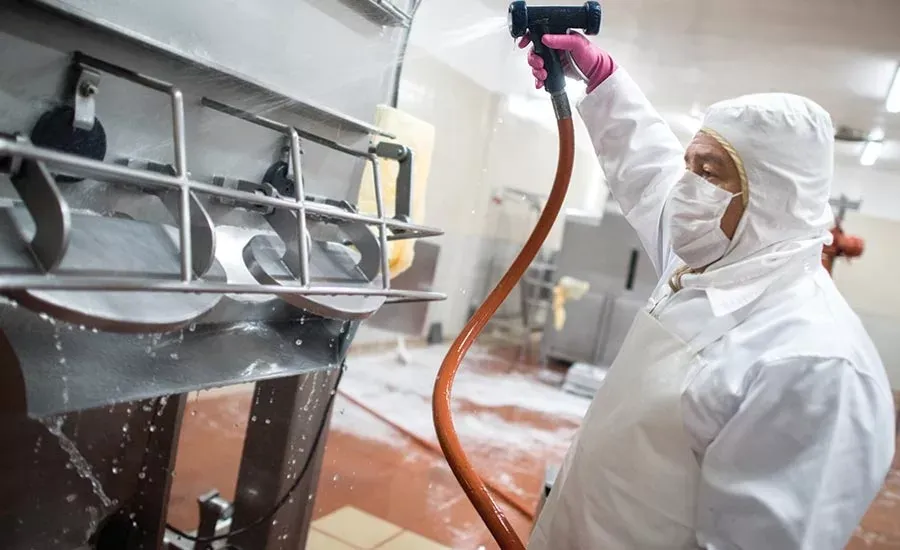 Worker in protective gear cleaning food factory equipment with a high-pressure hose spray.