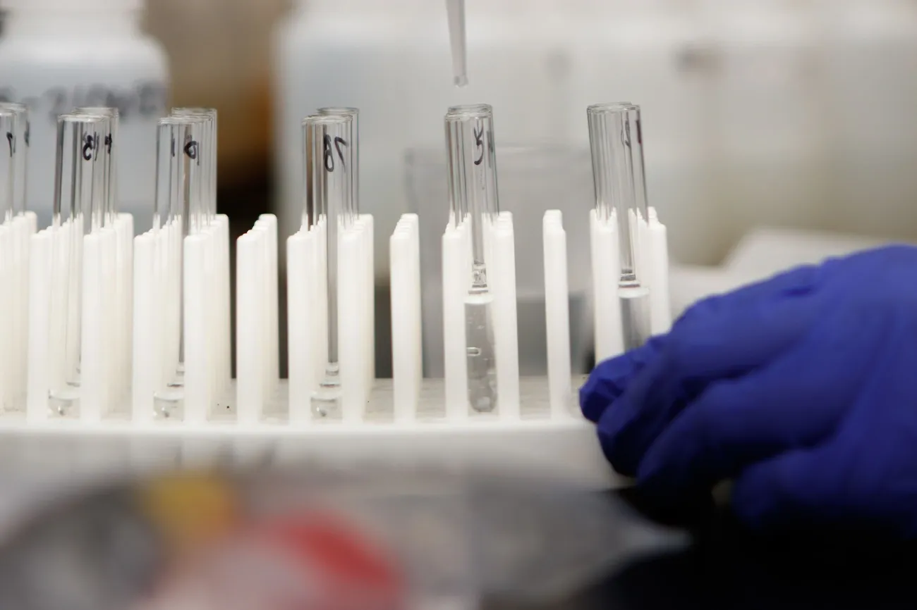 Gloved hand handling test tubes with liquid in a lab setting for food allergen control and testing.