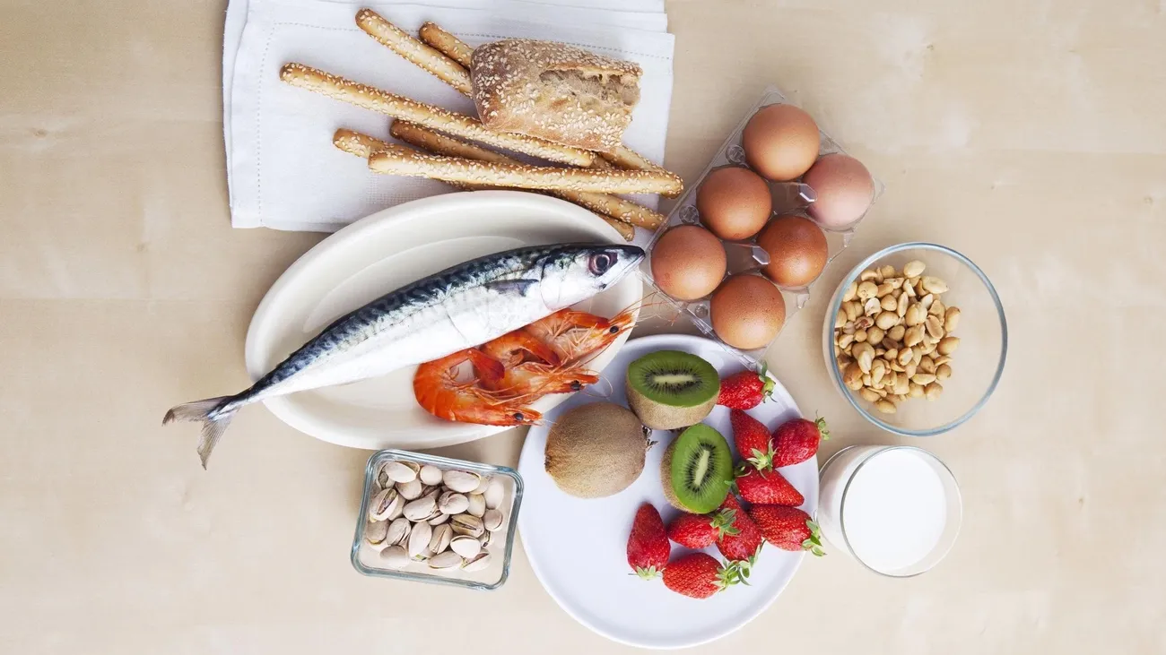 Various common food allergens including fish, shrimp, eggs, nuts, strawberries, kiwi, milk, bread, and breadsticks on a table.
