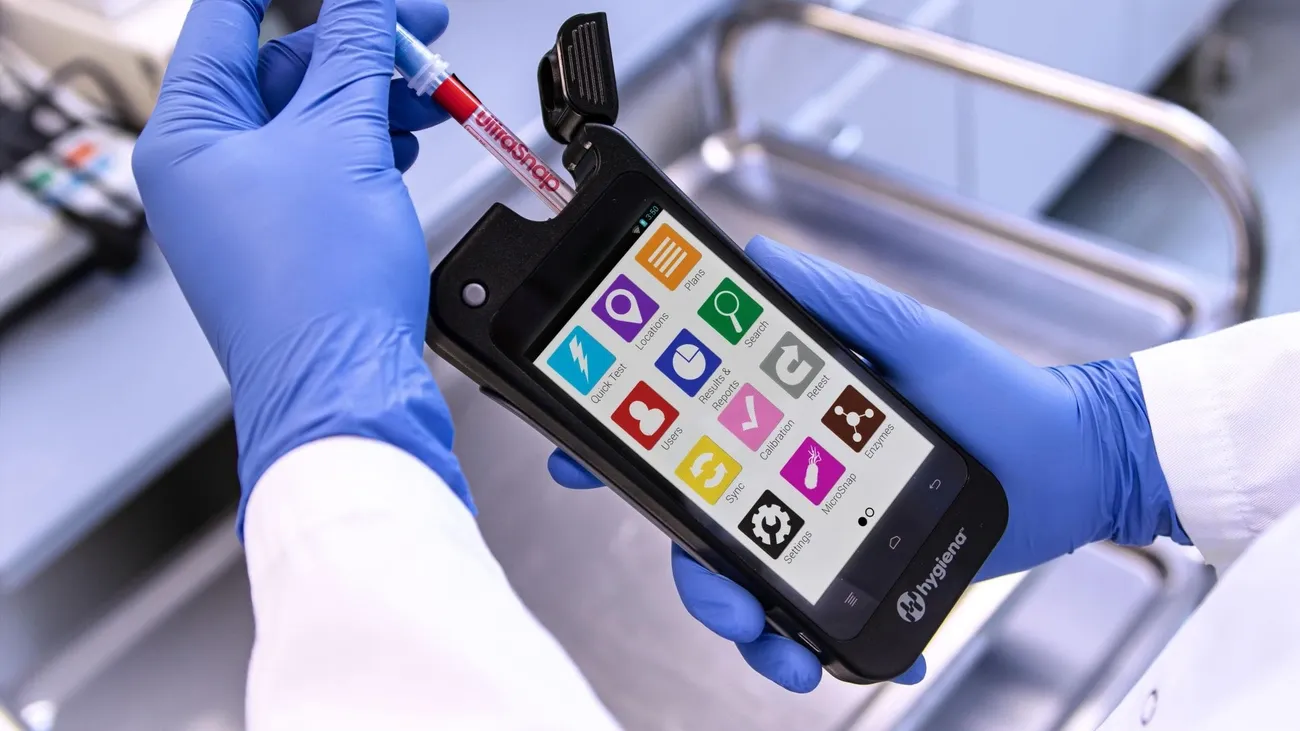 Hands in blue gloves holding a Hygiena device with a test swab inserted for ATP verification and monitoring.