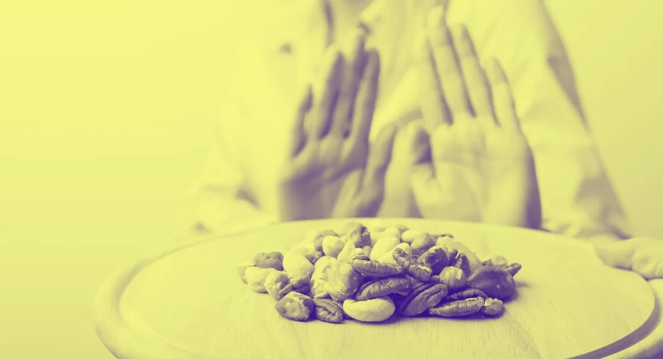 Person showing a stop gesture behind a pile of mixed nuts on a wooden board representing allergen control.