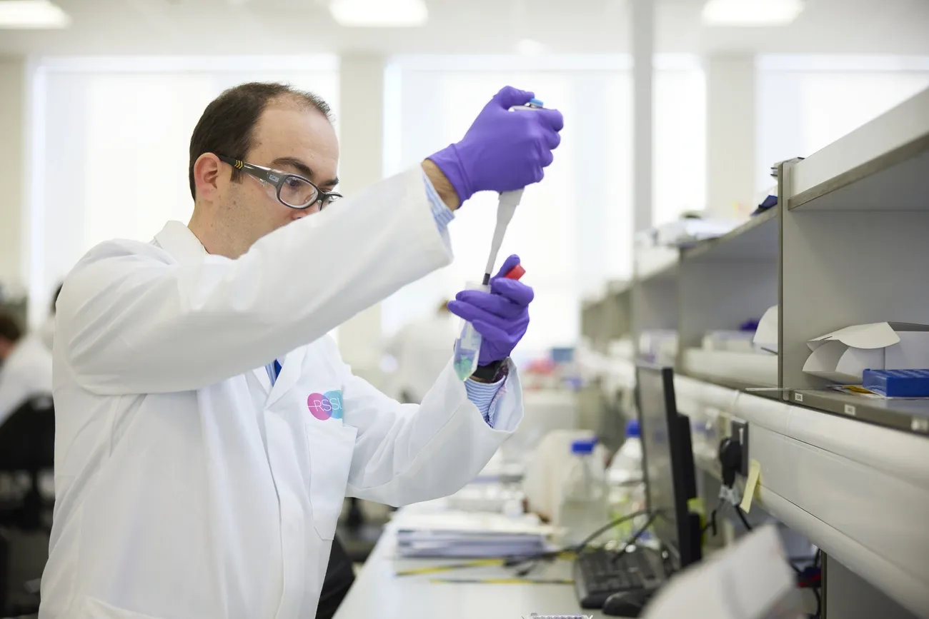 Scientist in a lab coat and purple gloves using a pipette to conduct food allergen testing in a laboratory.