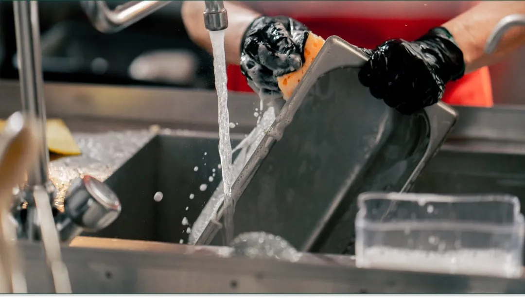 Person wearing black gloves washing a metal tray under running water in a kitchen sink