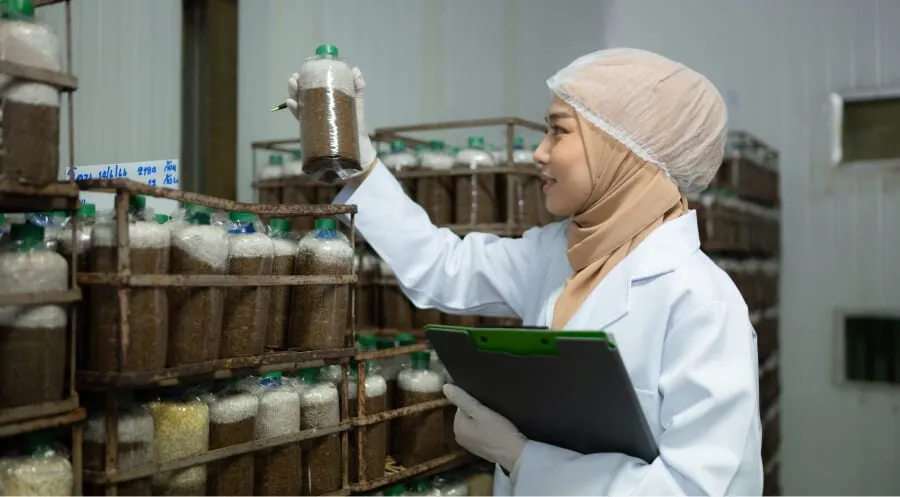 Person in protective clothing inspecting bottled samples in a storage facility with a clipboard in hand.