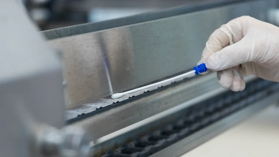 Gloved hand holding a swab for ATP testing on a metal surface in a lab or industrial setting.