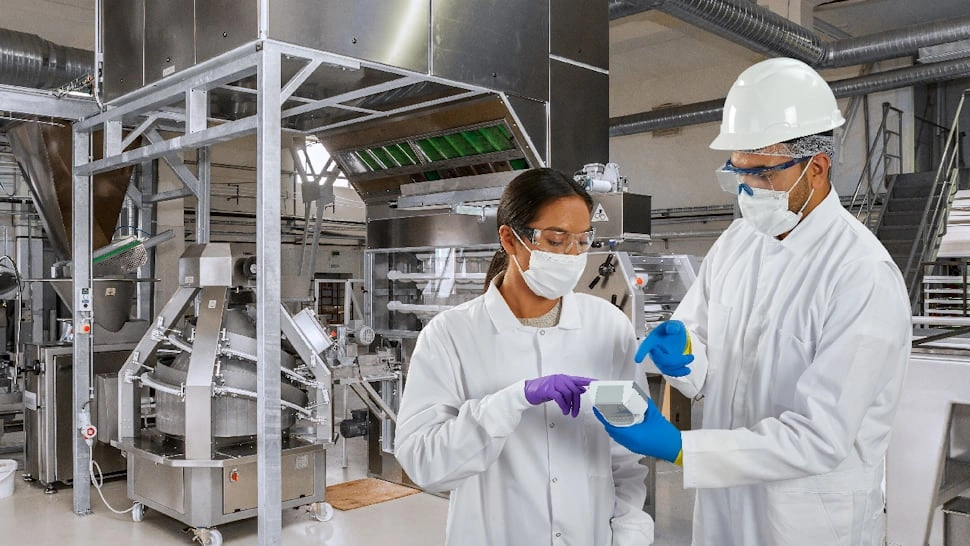 Two food industry workers in white lab coats and gloves conducting allergen testing in a factory setting.